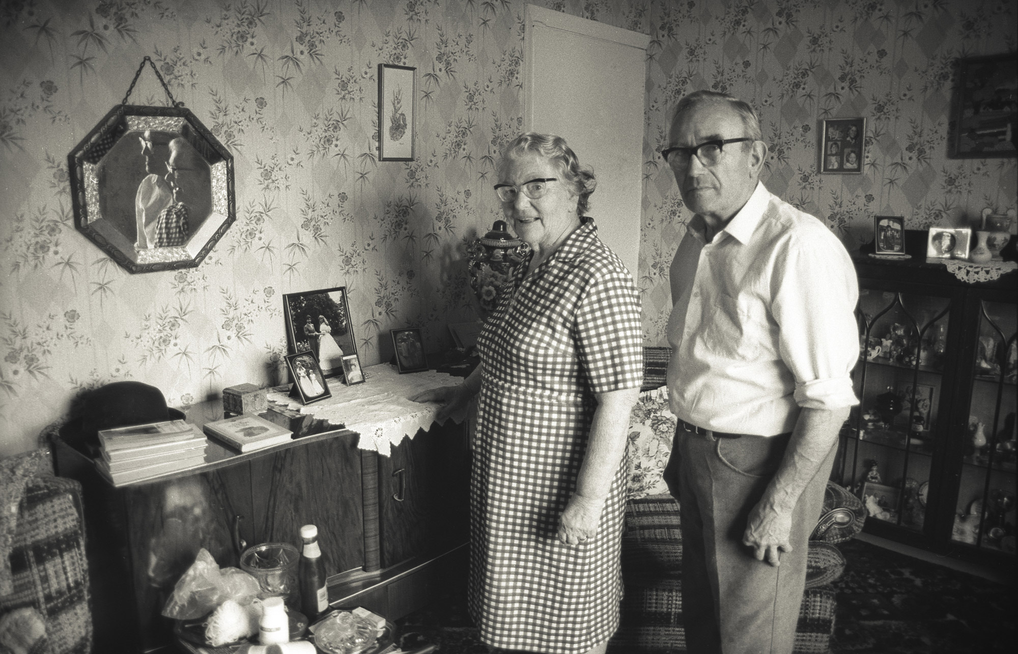 Beat and Fred standing together in their living room, surrounded by personal objects and photographs.