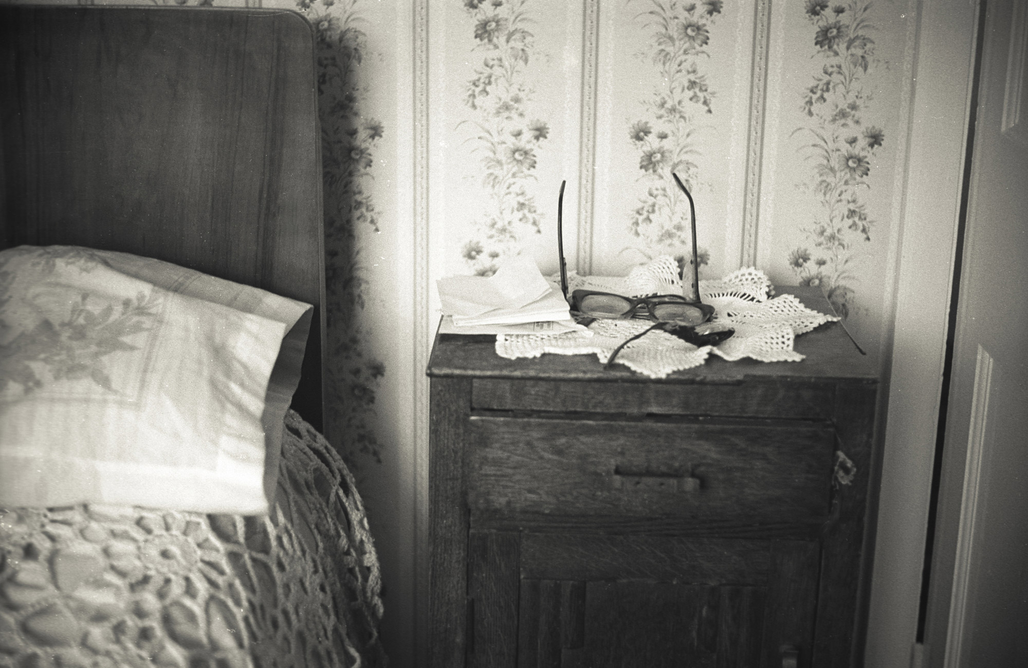 Black and white photograph of a bedside table with reading glasses, letters, and lace beside a bed.