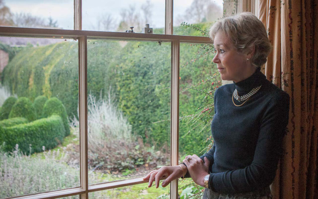 Woman standing by a window, looking out into a garden, softly lit and not retouched, photographed with restraint and honesty.