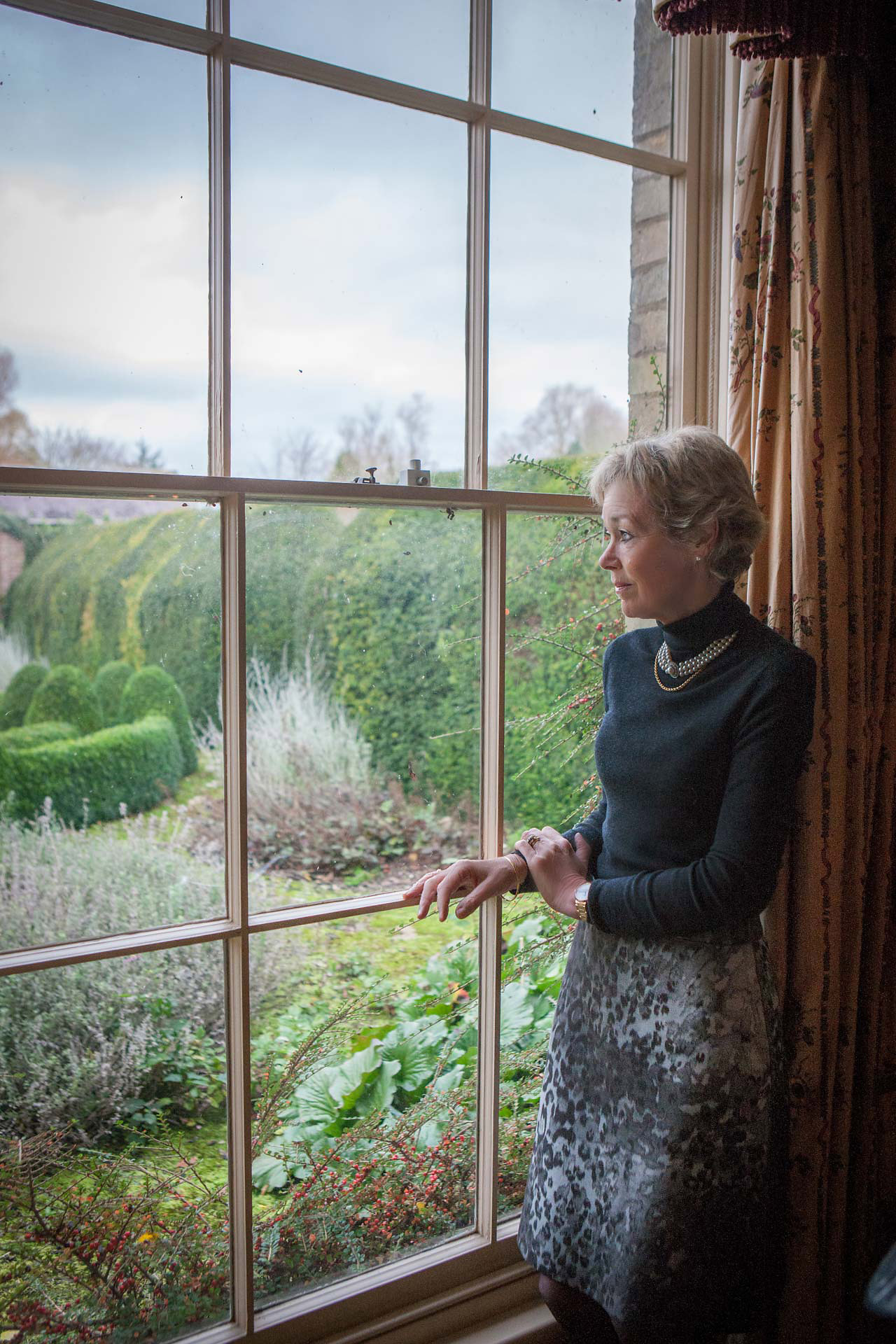 Woman standing by a window, looking out into a garden, softly lit and not retouched, photographed with restraint and honesty.
