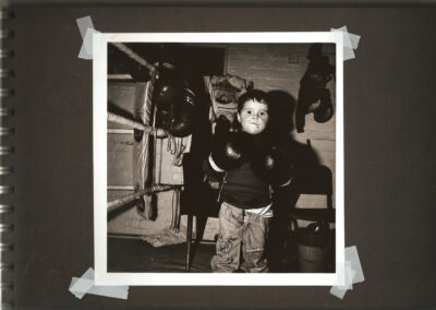 Young boy wearing boxing gloves stands inside a community boxing gym, looking toward the camera.