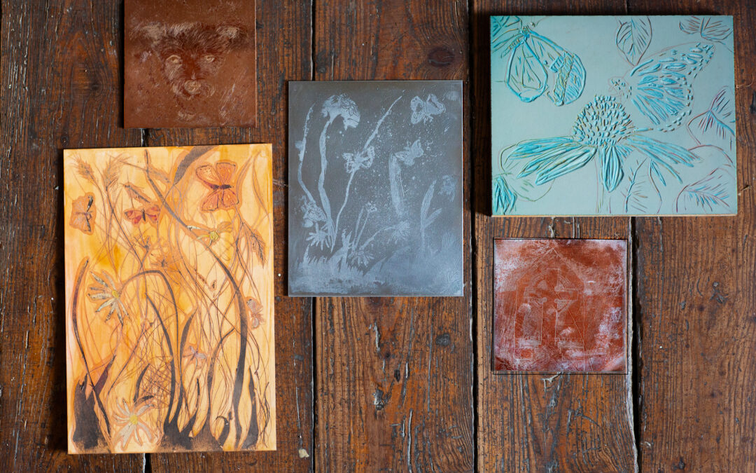 A photographed arrangement of experimental printmaking plates in copper, zinc, lino, and wood, showing early process marks and exploratory imagery laid out on a wooden surface.