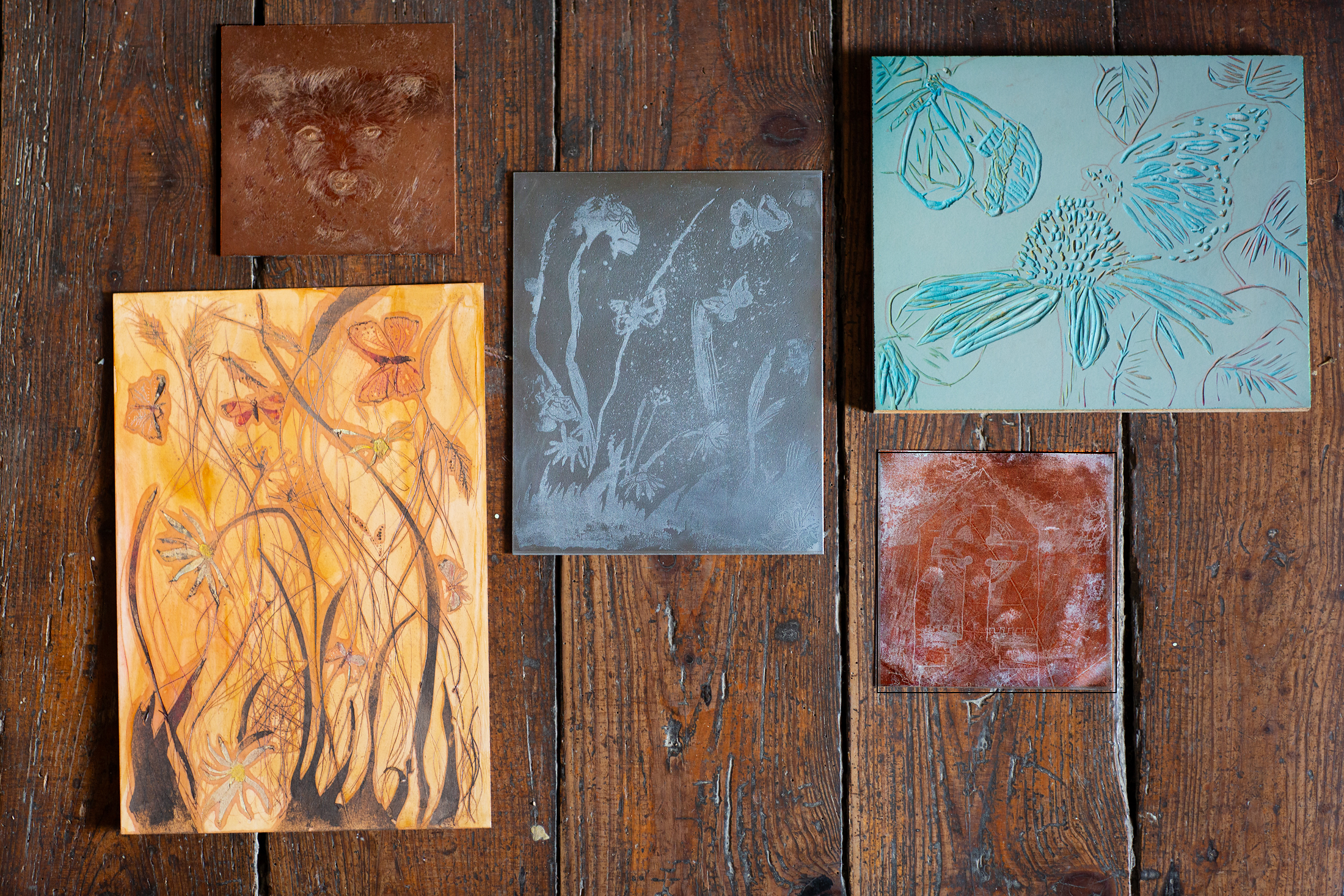 A photographed arrangement of experimental printmaking plates in copper, zinc, lino, and wood, showing early process marks and exploratory imagery laid out on a wooden surface.